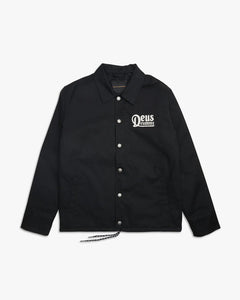 Wasserabweisendes Coach-Jacket von Deus in Schwarz mit silbernen Knöpfen. Auf der Brust ist eine kleine Stickerei in weiß "Deus Customs" angebracht.