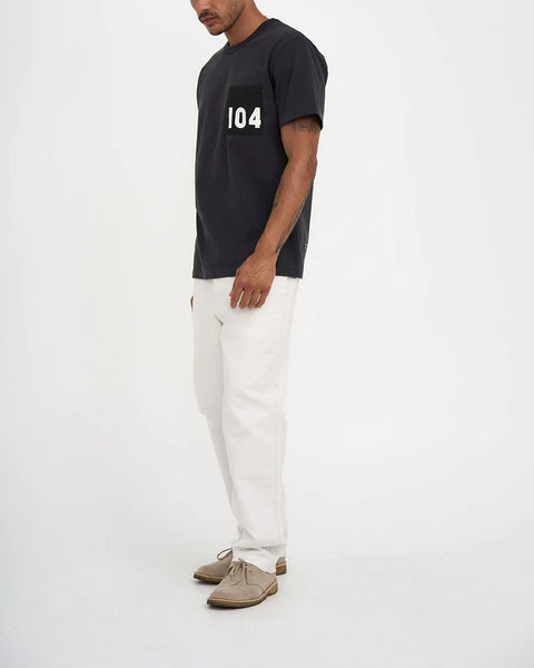 Locker geschnittenes T-Shirt von Deus in Anthrazit mit einer schwarzen Brusttasche. Ein männliches Model trägt das T-Shirt mit einer weißen Jeans.