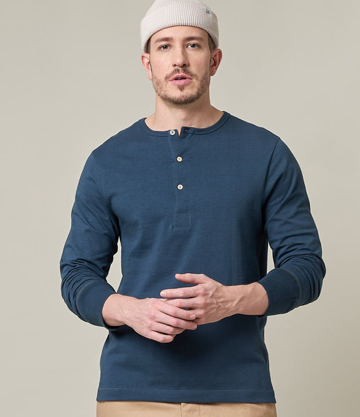 Merz b. Schwanen Henley in Langarm in der Farbe Mineral Blue, ein Mittelblau mit einer leichten Washed-Optik. Ein männliches Model trägt das Henley in Kombination mit einer beigen Chino und einem Beanie in Naturfarben.