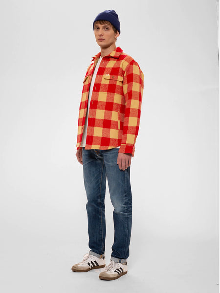Gefüttertes Overshirt von Nudie Jeans in Rot/Creme kariert. Ein männliches Model trägt das Hemd über einem weißen T-Shirt mit Beanie, verwaschenen Jeans und Sneakern. 
