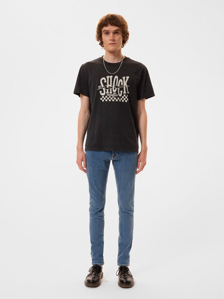 T-Shirt von Nudie Jeans in verwaschenem Schwarz, Print auf der Vorderseite "No Shock No Progress". Ein männliches Model trägt das T-Shirt mit einer schmalen hellblauen Jeans und schwarzen Lederschuhen.